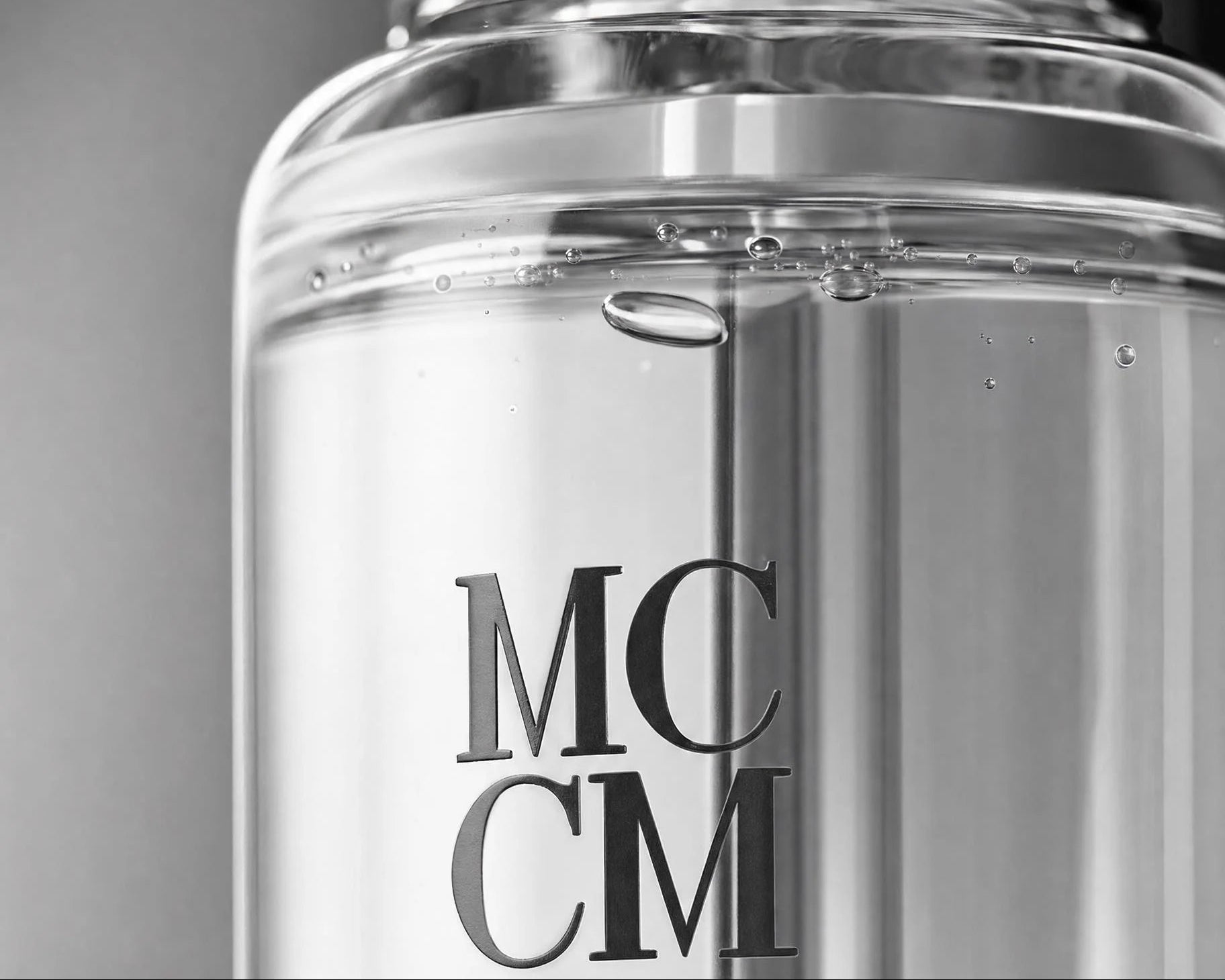 Close-up of a clear cosmetic bottle with a reflective silver cap and large dark lettering reading 'MCCM' on the front, shown against a soft gray background.